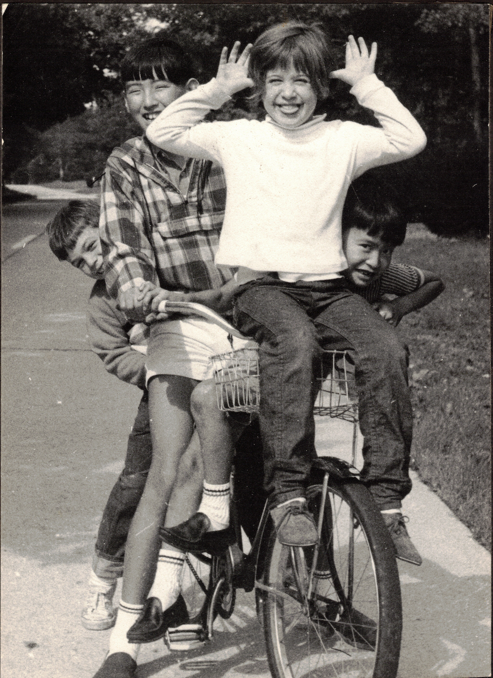 Black and white photo of one kid seated on a bike, another kid on the handle bars, and two other kiddos peeking out from either side. The kid on the handle bars holds their hands up next to the sides of her head, slightly sticking out her tongue with a grin.