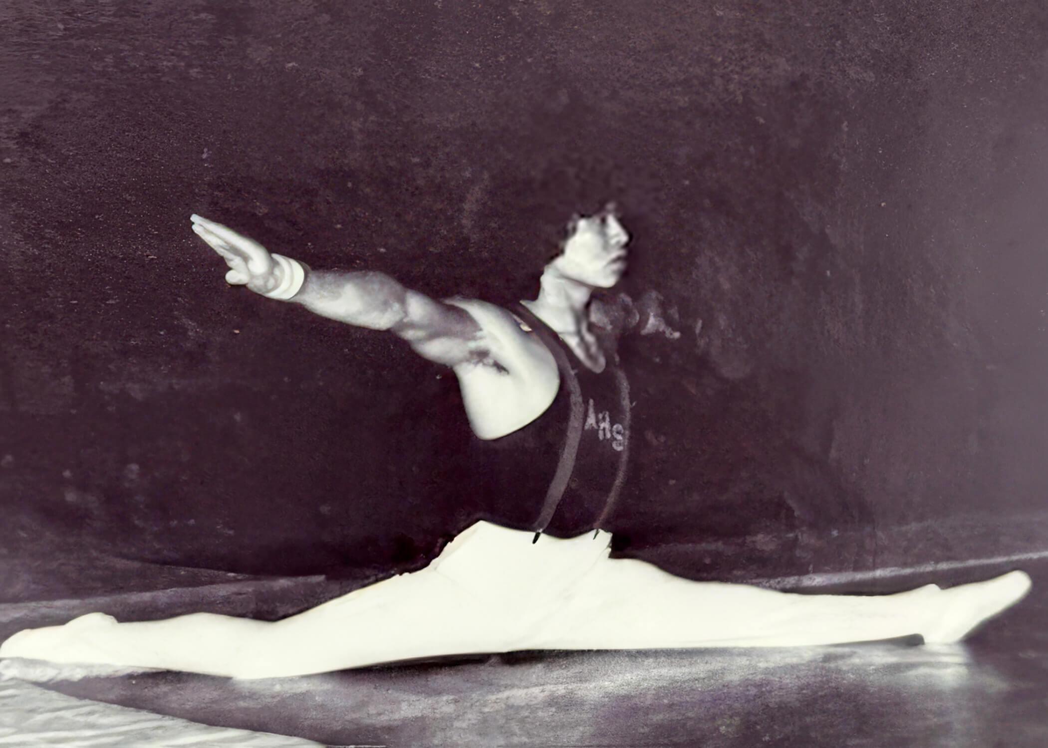 Rick in a full split on the gymnasium floor while holding his arms straight out to his sides, chest out. He gazes forward with confidence and focus.