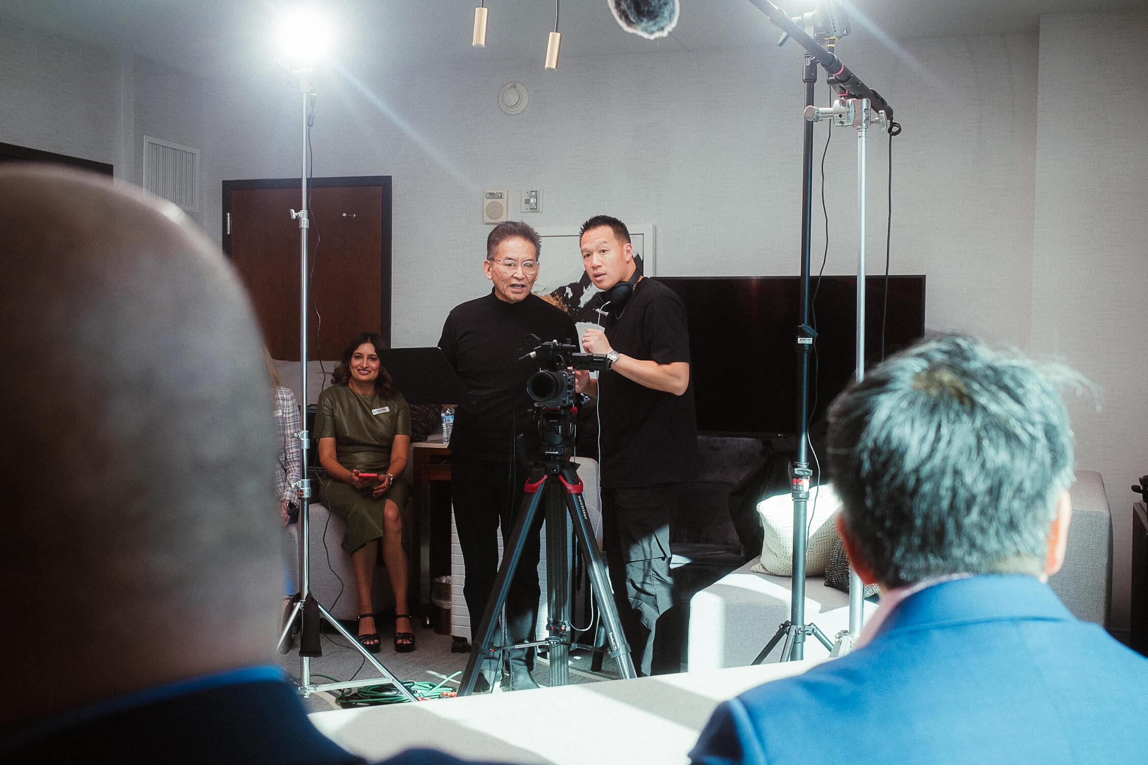 Rick and Austin surrounded by fancy lighting equipment stand behind a video camera on a tripod discussing the shot. Above them is a boom mic just out of frame.