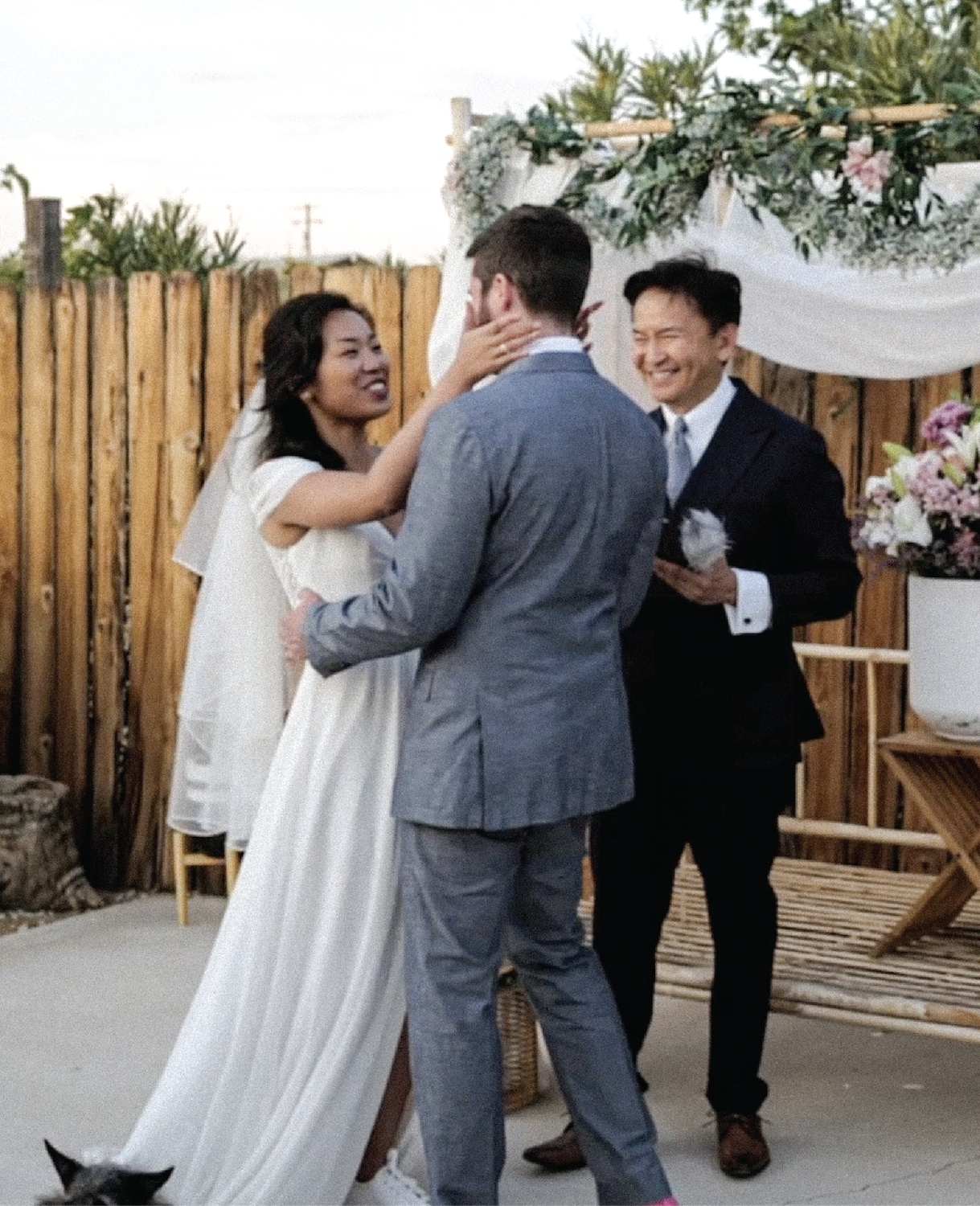 Aly is wearing a beautiful wedding dress. Ryan is decked out in a gray suit. They are facing one another, Rick behind them officiating the ceremony. Aly holds Ryan's face while he holds her hips as they prepare to share a celebratory kiss.